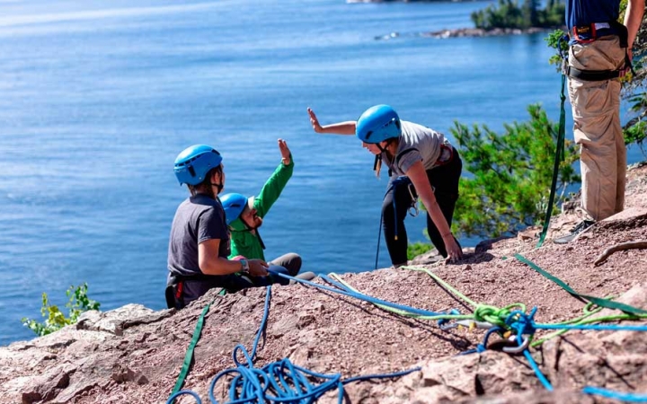 Two people wearing safety gear are secured by ropes as they high five near the edge of a cliff. There are two other people wearing safety gear on the cliff. They are high above a blue body of water.